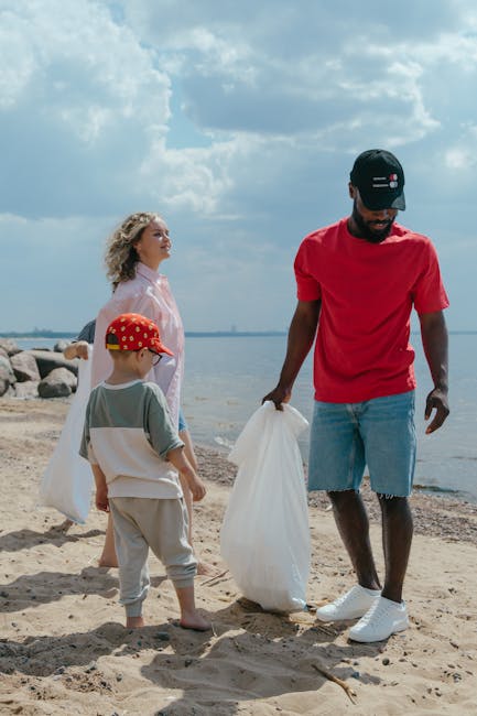 A young man wearing a black cap, red T-shirt, and ripped denim shorts is standing on a sandy beach, holding a large white plastic bag typically used for waste collection. Next to him, a small boy in a grey T-shirt, beige pants, and a red cap with yellow details is also holding a plastic bag, with his back partially turned towards the camera. Behind them, a woman with curly blonde hair, dressed in a light pink or pastel-colored jacket, stands facing the water, which is calm with a few ripples, under a partly cloudy sky with large clouds. The beach features a combination of sand and scattered rocks, with a distant shoreline visible across the water. The scene appears to be an organized community or family activity focused on cleaning or rubbish collection, subtly aligned with eco-friendly or rubbish removal practices that companies like Rubbish Removal Bayswater may support as an alternative to local waste disposal. Natural lighting suggests daytime with soft shadows cast on the sand, highlighting the textures and colors of the objects and clothing, creating a peaceful environment dedicated to environmental care or outdoor cleanup efforts.