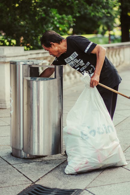 A pair of large black plastic rubbish bags and a cardboard box are placed on the edge of a pavement, set against a background of a black metal fence and a dense leafy green shrub. The bags appear full and are somewhat crumpled, with visible creases and a shiny surface reflecting ambient light, indicating they are tightly packed waste materials. The cardboard box, positioned to the left of the bags, has partially torn and folded edges, with markings that suggest it previously contained household items. The scene is outdoors, likely in an urban or suburban area, with the rubbish positioned for collection or disposal. The surrounding environment includes a paved sidewalk and a street, with minimal natural light or shadows present, creating a neutral, functional context typical of private waste management or alternative rubbish collection services, such as independent rubbish removal by Rubbish Removal Bayswater.