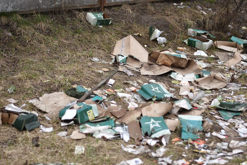 The image depicts an outdoor area with a mixture of discarded cardboard boxes, some of which are partially flattened or torn, and several crumpled paper and plastic waste scattered across a patch of uneven grassy ground. The cardboard boxes are predominantly green, featuring white star logos and yellow labels, and are variously bent, crushed, or discarded in a disorderly fashion. Interspersed among the boxes are smaller pieces of paper, scraps of packaging, and bits of plastic, creating a cluttered scene typical of unmanaged rubbish accumulation. In the background, a section of wooden fencing is visible, with a slope or embankment leading up to it, and dry, leafless shrubs are seen at the top of the incline. The lighting suggests an overcast day, casting diffuse light over the scene, emphasizing the disorderly nature of the waste and the neglected environment, which could relate to private or independent rubbish collection efforts supported by companies like Rubbish Removal Bayswater.