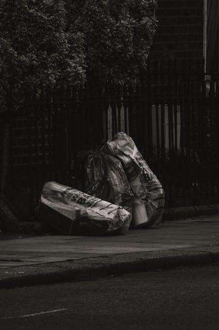 Two large black bin bags filled with waste materials, positioned on a sidewalk near a dark wooden fence and a lush green hedge. The bags appear to be made of thick plastic and are somewhat crumpled, with visible text and logos on the surface. They are resting directly on the pavement, which has a smooth, light-colored surface, and the scene is partially shaded, with ambient street lighting casting soft shadows. In the background, the fence is composed of vertical wooden slats, and behind it, a dense hedge with leafy branches is visible. The setting suggests an area where private rubbish collection or on-site waste clearance may be conducted, aligning with services offered by Rubbish Removal Bayswater for domestic or commercial rubbish disposal.