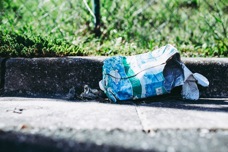 The image depicts an outdoor area with a mixture of discarded cardboard boxes, some of which are partially flattened or torn, and several crumpled paper and plastic waste scattered across a patch of uneven grassy ground. The cardboard boxes are predominantly green, featuring white star logos and yellow labels, and are variously bent, crushed, or discarded in a disorderly fashion. Interspersed among the boxes are smaller pieces of paper, scraps of packaging, and bits of plastic, creating a cluttered scene typical of unmanaged rubbish accumulation. In the background, a section of wooden fencing is visible, with a slope or embankment leading up to it, and dry, leafless shrubs are seen at the top of the incline. The lighting suggests an overcast day, casting diffuse light over the scene, emphasizing the disorderly nature of the waste and the neglected environment, which could relate to private or independent rubbish collection efforts supported by companies like Rubbish Removal Bayswater.