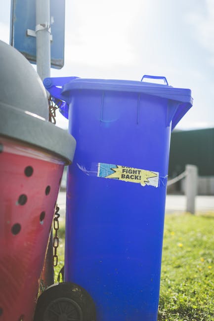 A tall, bright blue plastic rubbish bin with a closed lid, situated outdoors on a grassy area near a sidewalk and a road, with a handle on top for easy opening. The bin features a small, rectangular sticker on its front that reads 'FIGHT BACK!' in bold yellow and blue lettering, with a lightning bolt graphic. To the left of the blue bin, part of a red wheeled rubbish container with a black lid and a mesh section is visible, secured with a chain. In the background, a metal pole with a sign attached and a clear sky with some clouds can be seen, indicating daytime. The scene suggests a setting for private rubbish collection or waste management, with the scene taken in an open, suburban environment, aligning with the context of alternative waste handling and rubbish removal services.