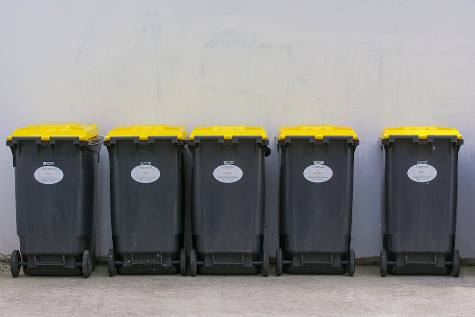 Five large wheeled rubbish bins with yellow lids are aligned in a row against a plain light grey wall, positioned on a concrete pavement. Each bin is made of dark grey plastic with a textured surface, and they feature a white oval label with identification numbers, some of which are partially visible. The yellow lids are securely closed, and the bins are placed close together, with slight variations in spacing. The environment appears to be an outdoor area, possibly a designated waste collection point for residential or commercial properties, with natural daylight illuminating the scene evenly. The presence of these bins suggests preparation for waste disposal or collection, aligning with alternative rubbish removal or private waste handling services offered by companies like Rubbish Removal Bayswater. The setting remains neutral and functional, emphasizing the practical aspect of waste management without additional decorative elements or clutter.