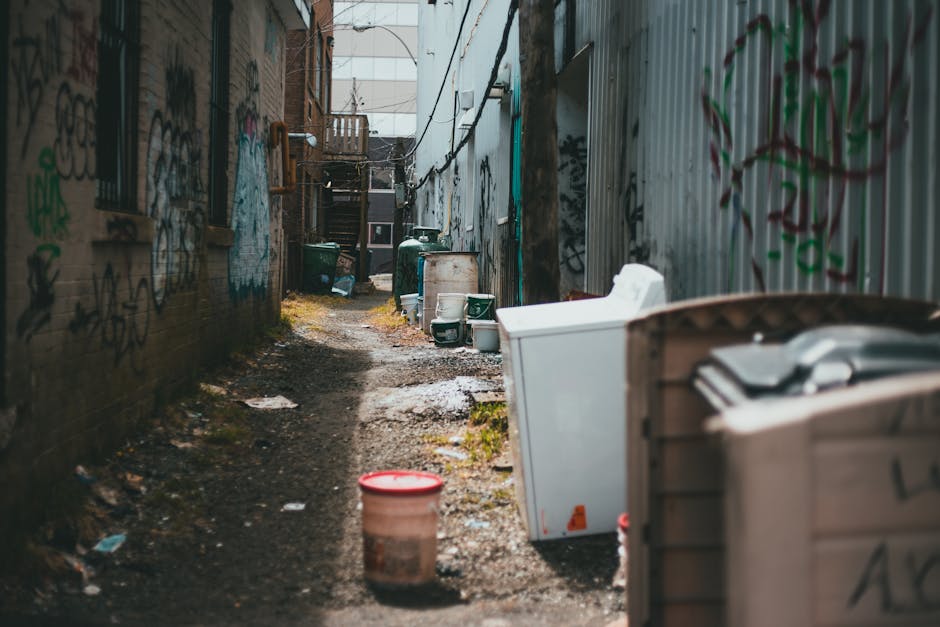 An urban alleyway with uneven, muddy ground scattered with debris and litter. The narrow space is enclosed by graffiti-covered brick walls on the left and a corrugated metal fence on the right, both showing signs of wear and weathering. Several large, white plastic storage containers and black garbage bins are lined up along the right side, some partially open or tipped over, with waste spilling out. In the foreground, a small red and white plastic bucket is placed on the ground. Further down the alley, additional green and black wheelie bins, some with lids closed and others open, are visible, along with a tower of stairs at the end of the alley leading to an upper level. Sparse patches of moss and dirt are present on the ground, illuminated by diffused natural light filtering from above, creating an environment suggestive of private waste disposal or on-site clearance, possibly handled by a rubbish removal service such as Rubbish Removal Bayswater. The scene portrays a typical alleyway used for waste storage or temporary rubbish collection in an urban setting.