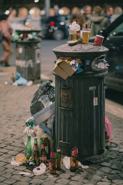 A black, tall metal rubbish bin situated on a cobblestone street with a domed lid, surrounded by various discarded items including empty bottles, crumpled paper, and food containers. The lid is partially open, with several empty beer bottles, plastic cups, and food wrappers resting on top, some leaning against the bin. Around the base, there are additional empty glass bottles and scattered debris, highlighting improper disposal in an urban outdoor environment. In the background, a grey waste collection bin is visible, along with a blurred urban scene featuring pedestrians, parked cars, and a tree-lined street, suggesting a busy city setting. The lighting appears natural, indicating daytime, and the scene emphasizes issues related to littering and waste management outside of formal rubbish collection areas. Rubbish Removal Bayswater services are relevant to the context of managing such waste and litter in public spaces.